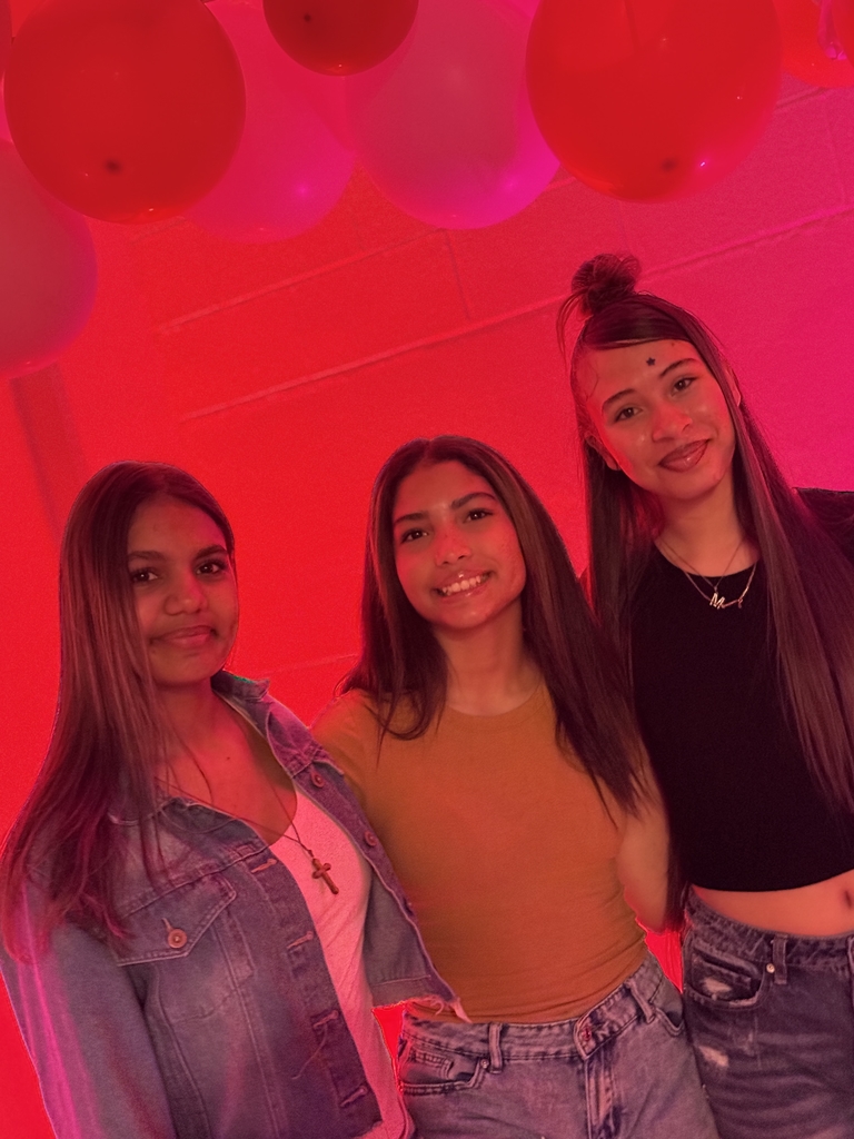 Three students smile together in front of a valentine day backdrop.