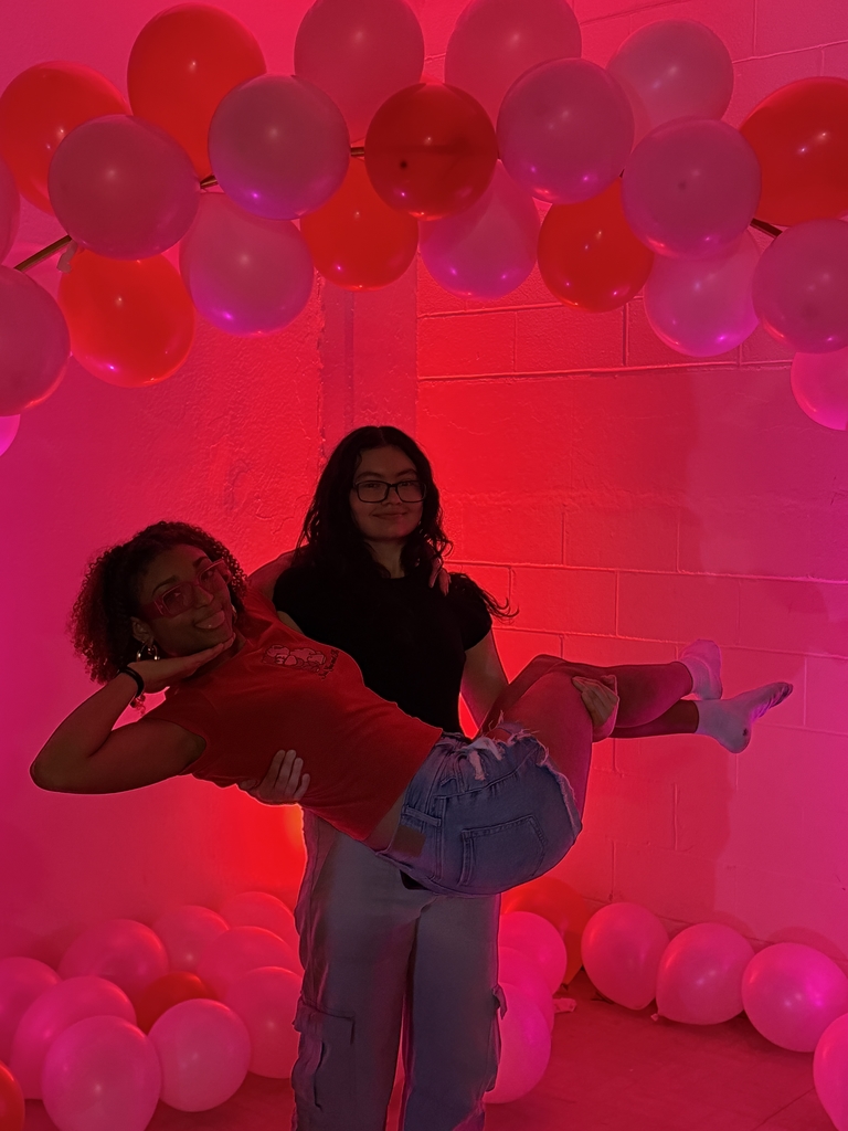 Two students smile together in front of a red backdrop.