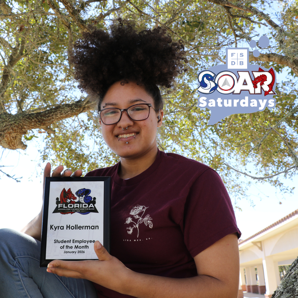 Kyra is wearing a maroon shirt with a small white outline of a plant with her hair up and is wearing black frame glasses. She is holding up a plaque that says "Kyra Hollerman Student Employee of the Month January 2026"