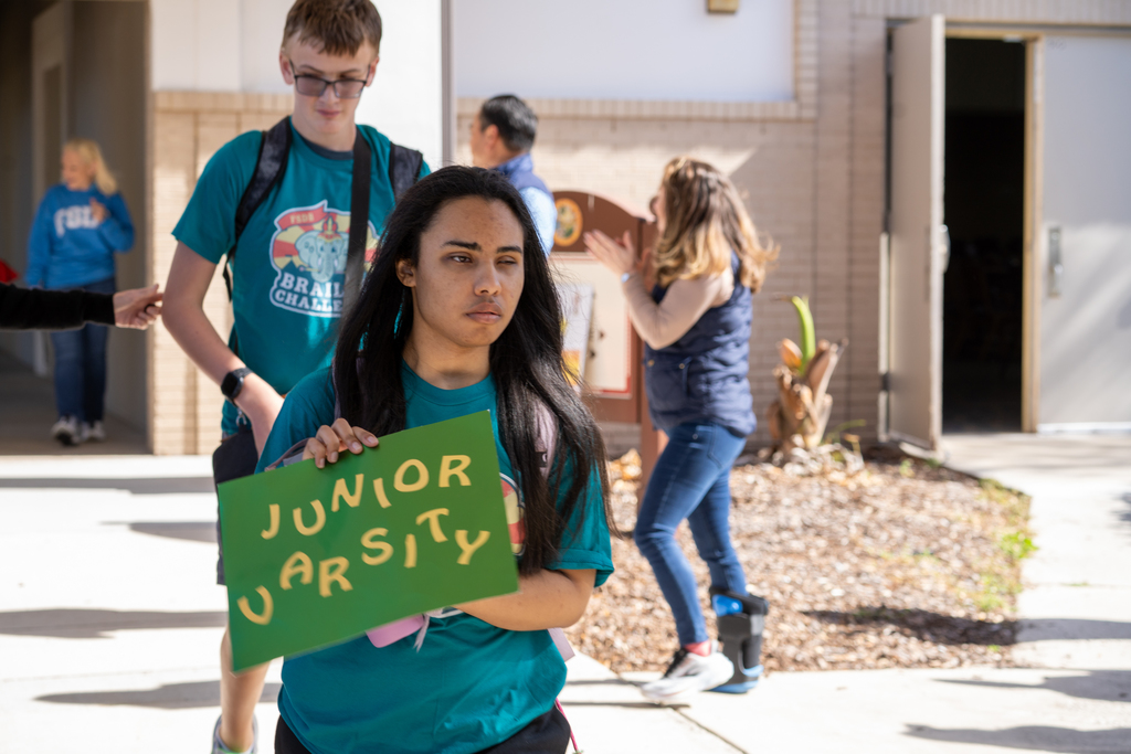 A student wearing a teal color shirt holds a green sign that says "Junior Varsity" as she walks through the crowd outside. 