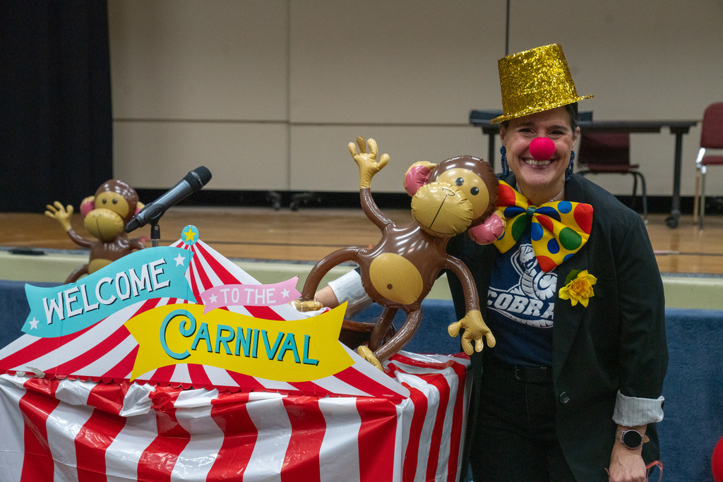 Tracie Snow is dressed like a clown smiling with a monkey blow up and podium covered in a red and white tent with a "Welcome to the Carnival " inside the music building. 