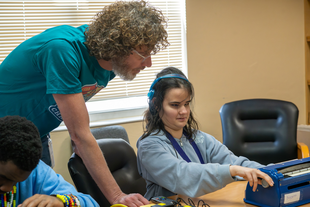 A student is sitting with a teacher over her shoulder telling her the rules inside a classroom. 