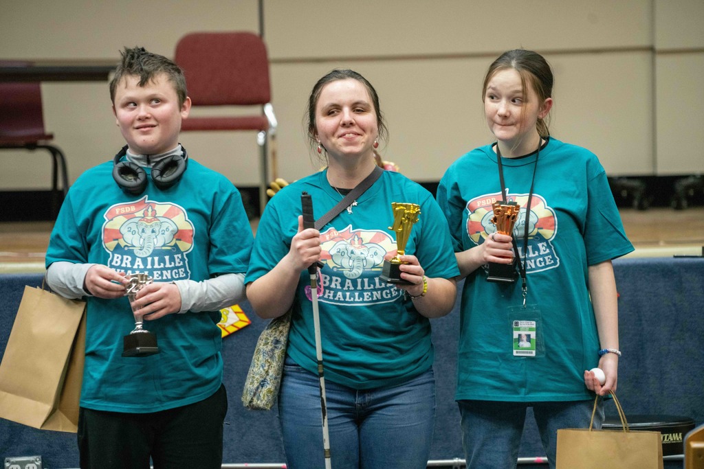 Three students wearing matching color team shirts with the circus Braille Challenge logo holding their trophies smiling for the camera inside the Music Building. 