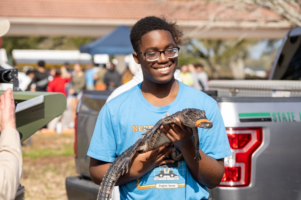 A student holding a baby alligator and smiles for a photo outside. 