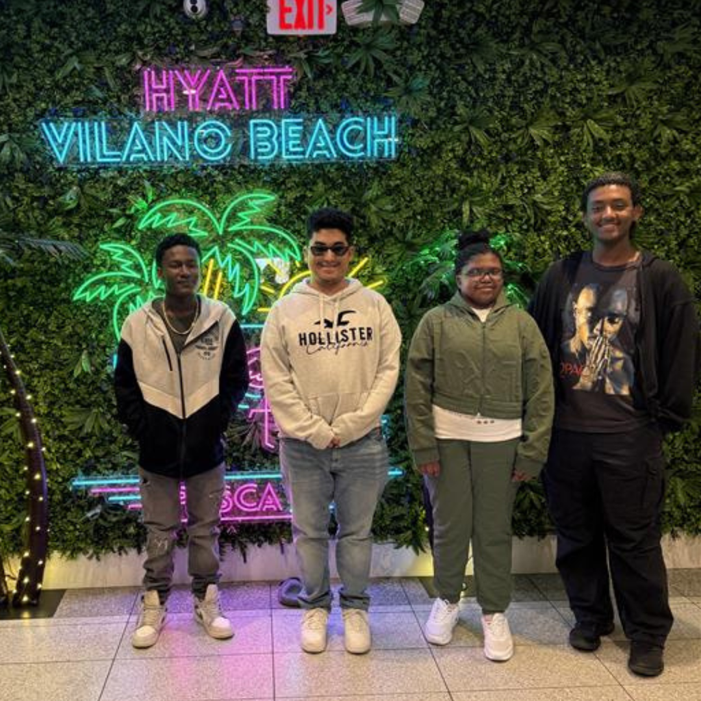 Group of students smile by a grass wall with led lights that say "Hyatt Vilano Beach" . 