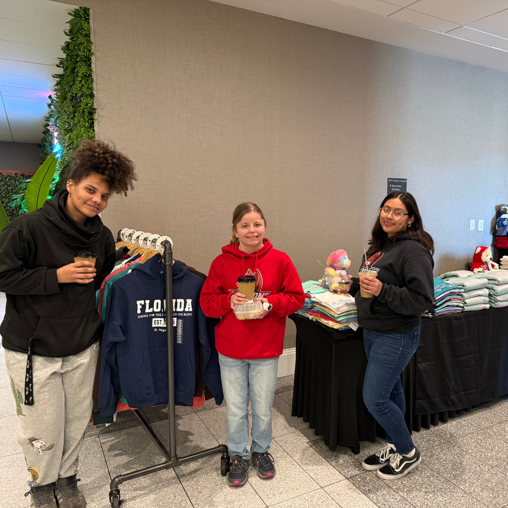 Group of students smile with their coffee and treats by the DB Designs merch inside the hotel building.