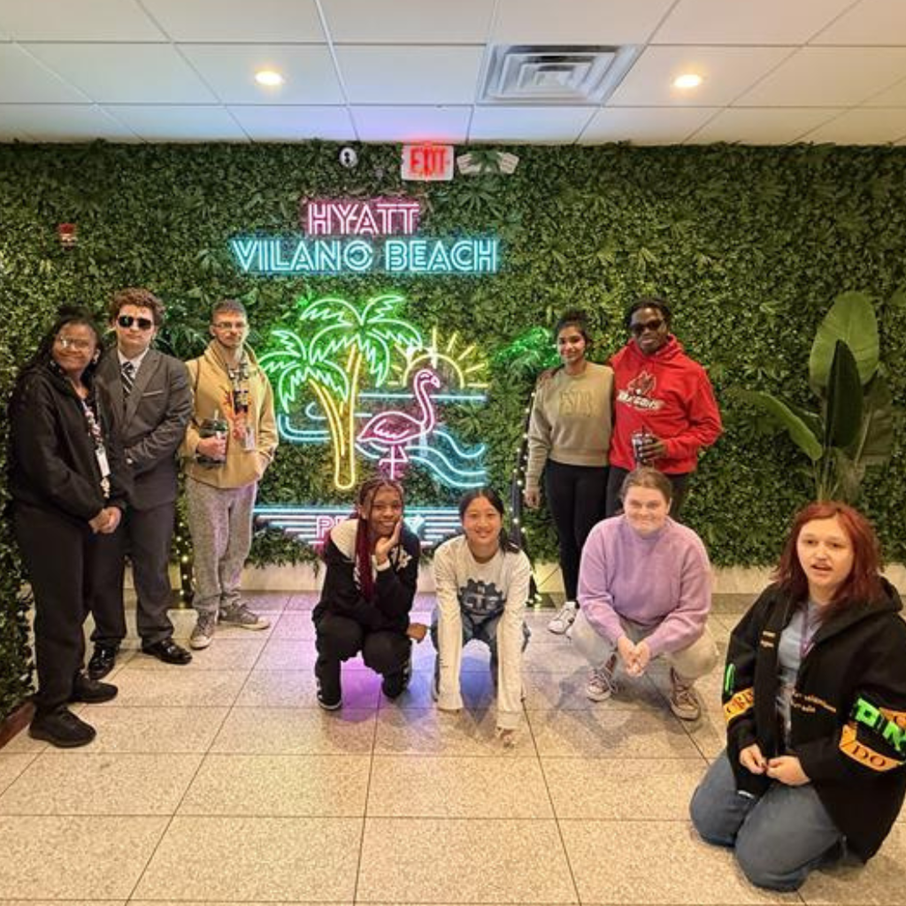 Group of students smile by the grass wall and led lights of a palm tree, sun, and flamingo and the wording "Hyatt Vilano Beach". 