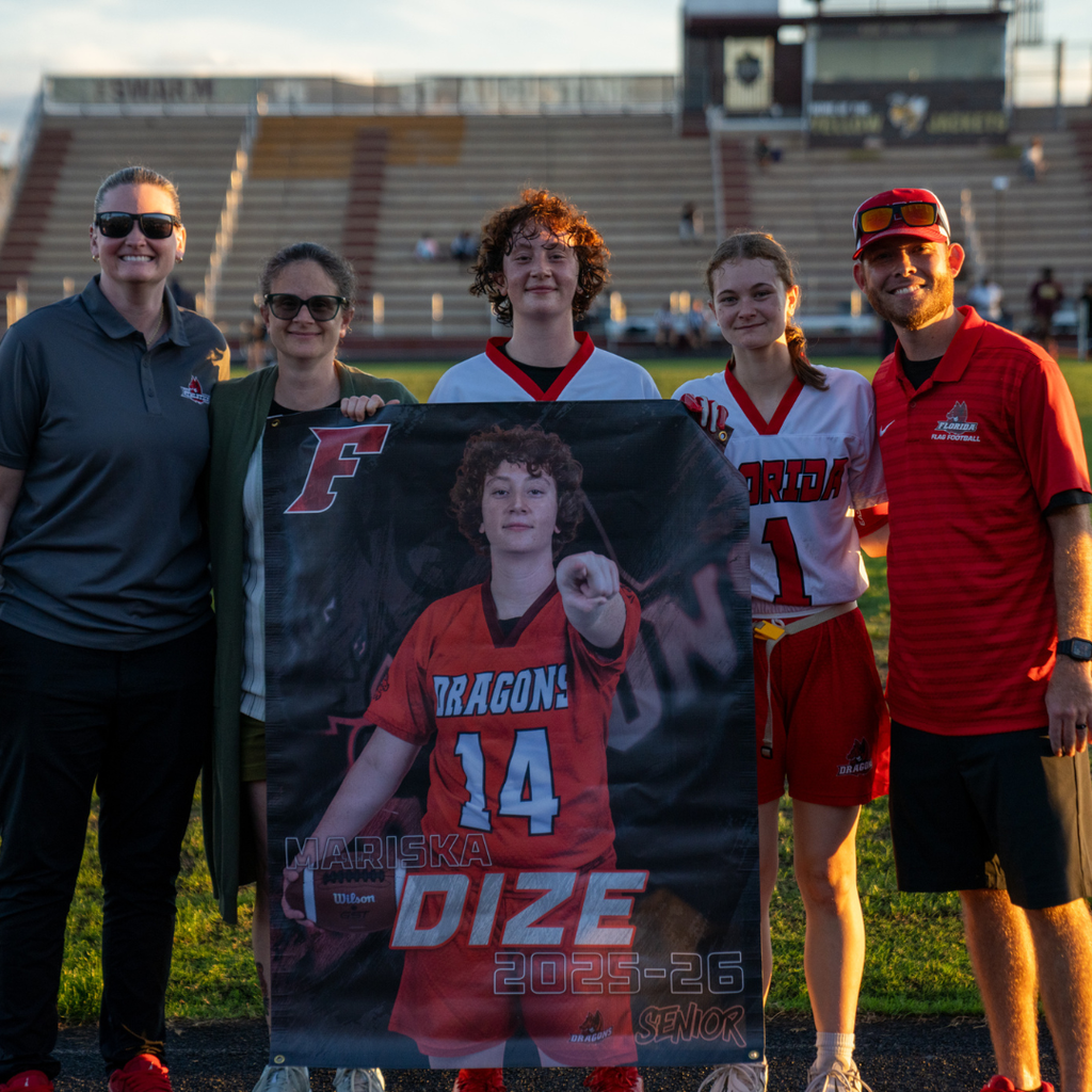 A senior holds her banner and smiles with her family and coaches on the field.