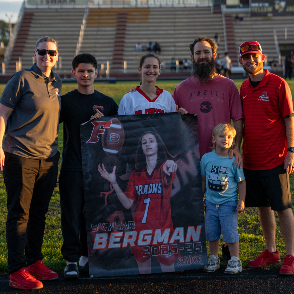 A senior holds her banner and smiles with her family and coaches on the field.