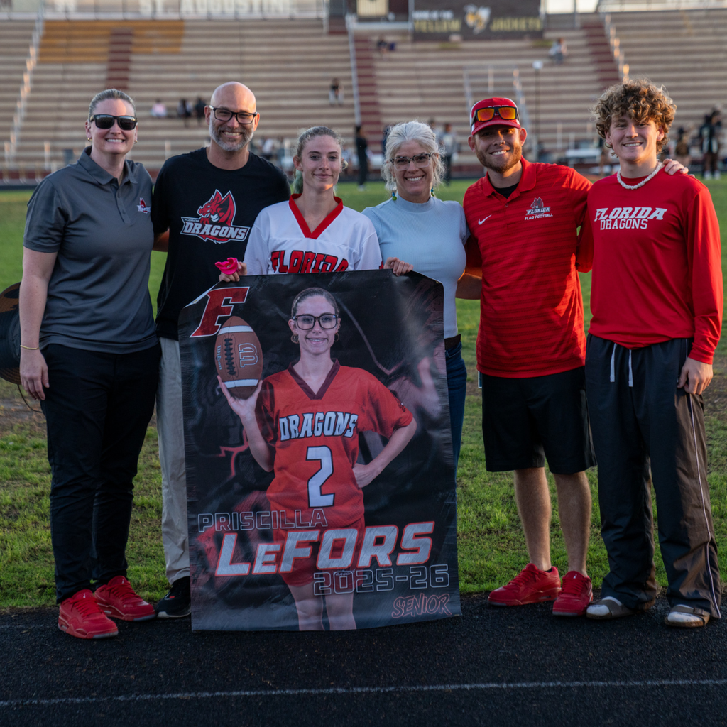 A senior holds her banner and smiles with her family and coaches on the field.
