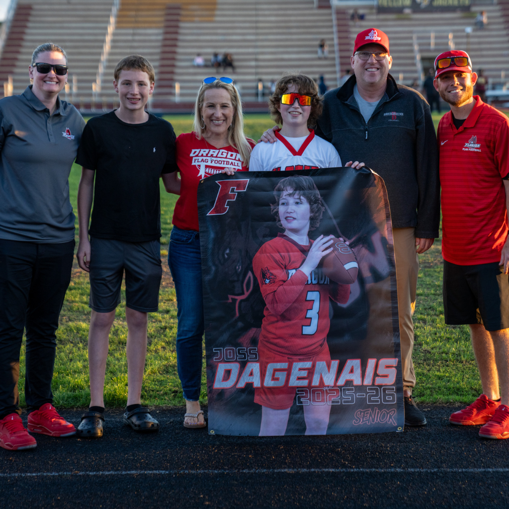 A senior holds her banner and smiles with her family and coaches on the field.