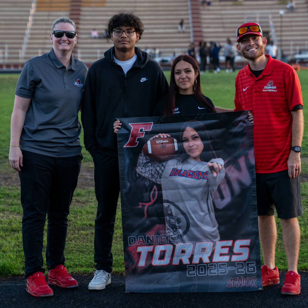 A senior holds her banner and smiles with her friend and coaches on the field.