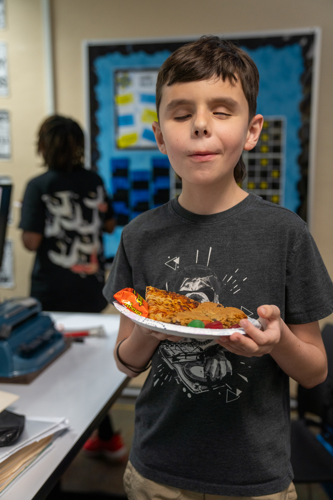 A student smiles with his pizza, brownies and candy inside the classroom. 