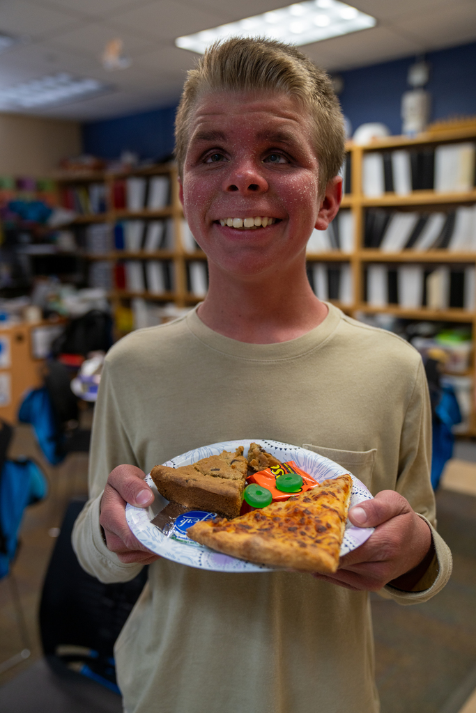 A student smiles with his pizza, brownies and candy inside the classroom.