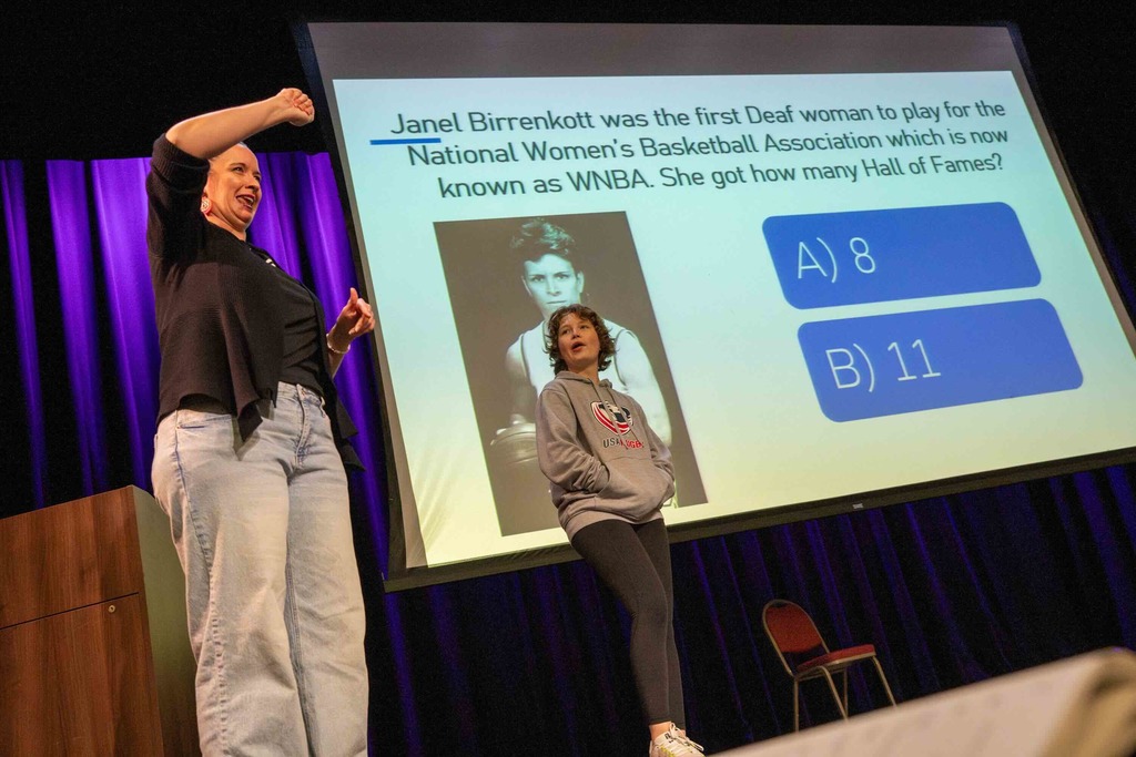 Dr. Renca signs on stage with a deaf high school student and an A or B question on the screen behind her on stage in Kirk.