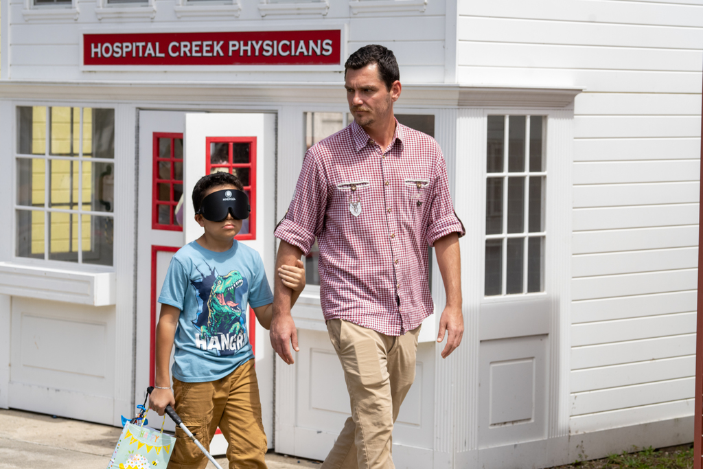 A staff member help guide a student who is wearing eye mask looking for eggs outside of Kids Town.