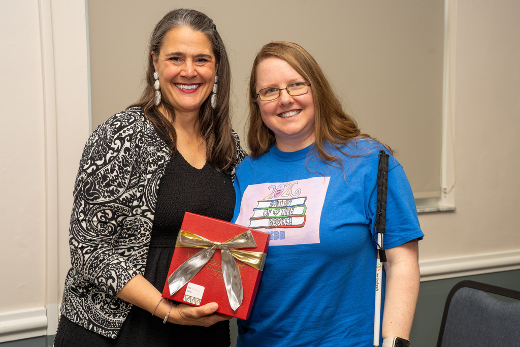 President Tracie Snow holds a red gift box with the BEMS Teacher of the Year who is wearing a blue colored shirt, smiles for her photo in CLD.