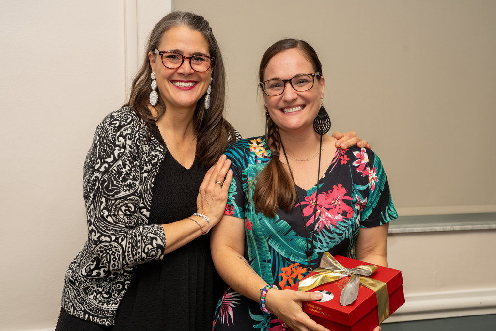 President Tracie Snow is smiling with the DES Teacher of the Year who is wearing a Hawaiian flower dress and holds a red gift box inside CLD.