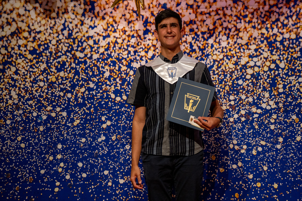 Damian holds his NHS booklet and smiles on stage with a blue and gold sparkle backdrop.