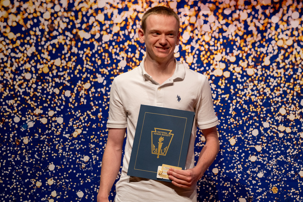 Tyler smiles and holds his NHS booklet on stage with a blue and gold sparkle backdrop.