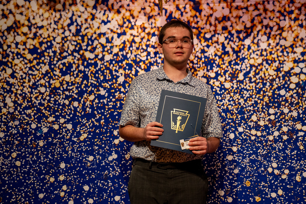 Gordon smiles with his NHS booklet with a blue and gold sparkle background inside the music building.