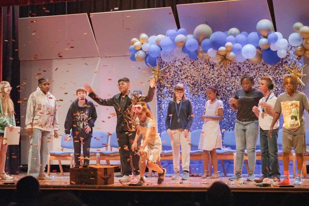 A group of students on stage in the Music Building with gold confetti scattered and blue and gold balloons on the top of the stage.