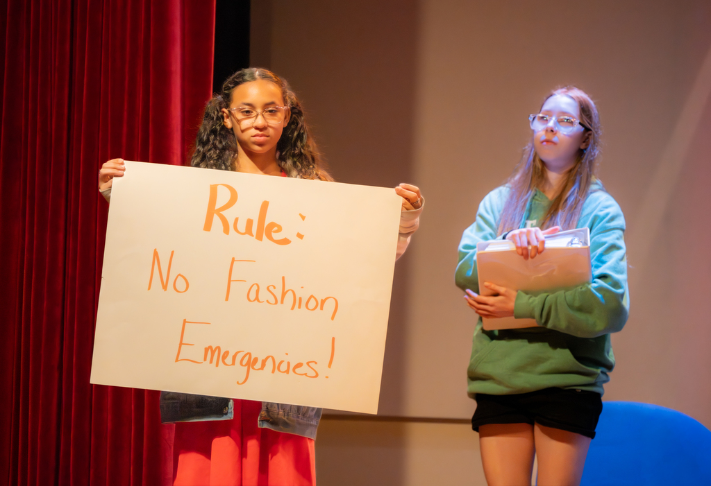 One student is holding a white sign that says "Rule: No Fashion Emergencies!" and the other student holds a big binder acting on stage in the Music Building.