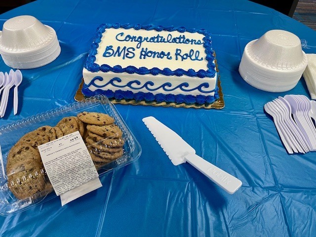 Blue table cloth with a cake that is written in blue says "Congratulations BMS Honor Roll" with cookies and plastic plates, forks and bowls. 