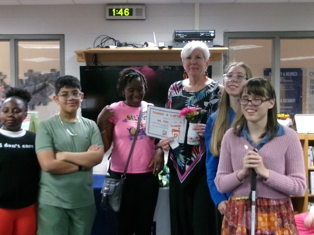 Students smile with Ms. Fertsch holding her flowers and her own certificate for National Assistant Principal Week inside the library.