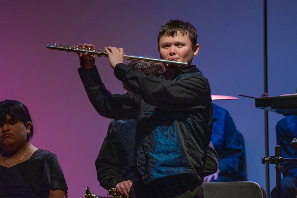 A Blind FSDB student wearing a black long sleeve with blue vest stands on stage in Music Building and performing his flute. 