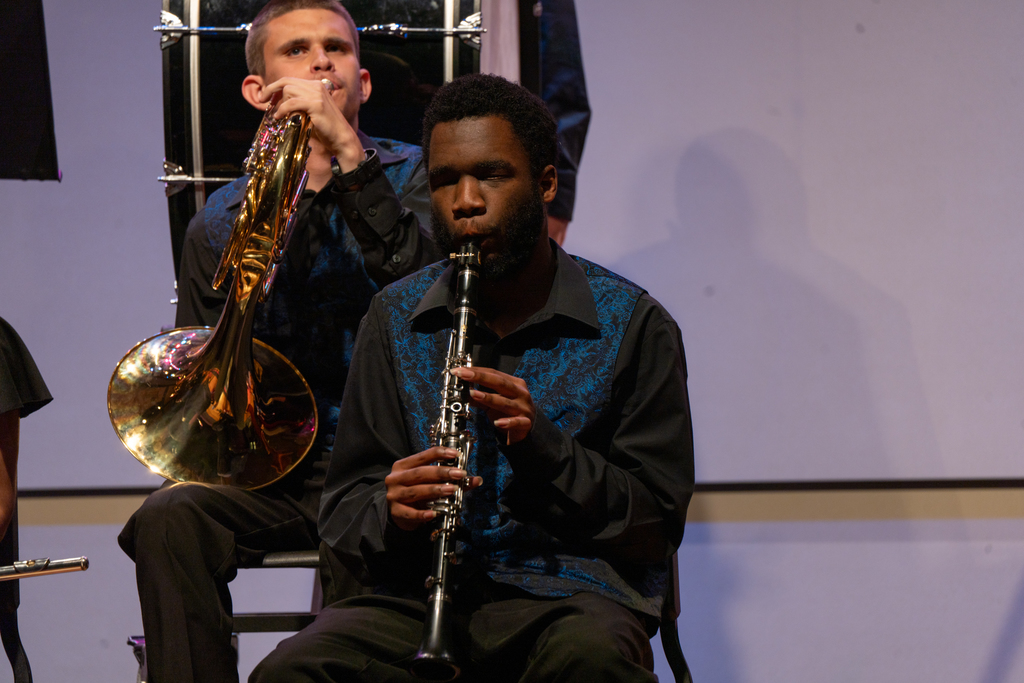 A FSDB student is wearing a black long sleeve shirt and blue vest sitting on a black chair in the Music Building performing his clarinet. 