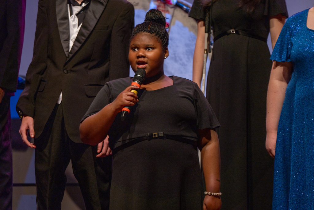 A Blind FSDB student is wearing a black concert dress holding the microphone and singing on stage in the Music Building. 