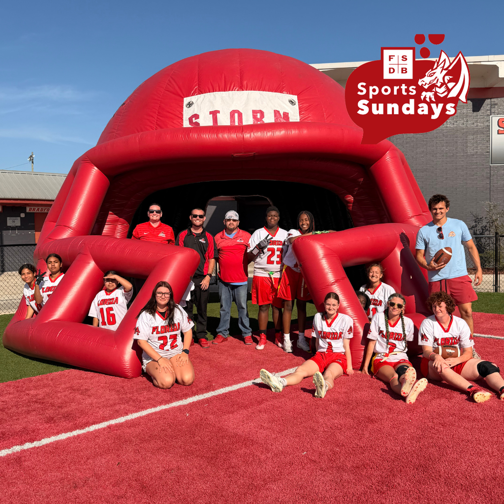 Varsity Girls Flag Football team smile for a photo on a red turf and a big red blow up helmet that says "Storm" on the top. 