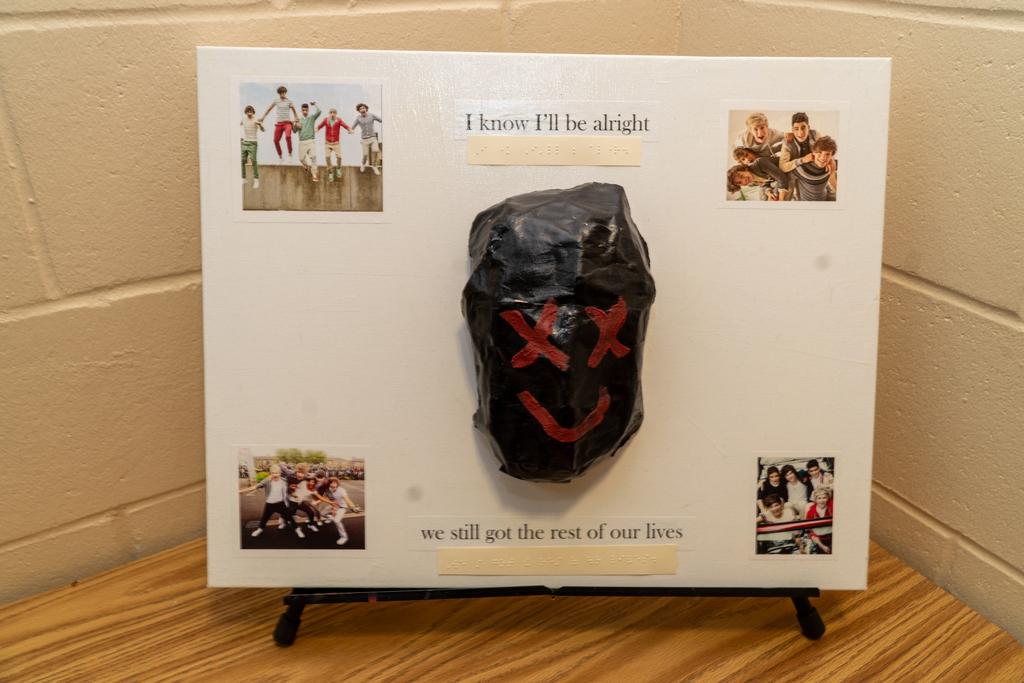 Creation by a blind student of a white canvas board with photos of One  Direction band members on all four corners, with a black mask with red eyes and mouth, along with the phrase "I know I'll be alright, we still got the rest of lives."