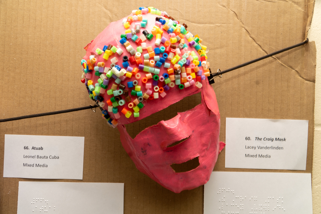 Creation of a mask that is pink and covered with colorful beads created by a Blind student held for display on a cardboard. 