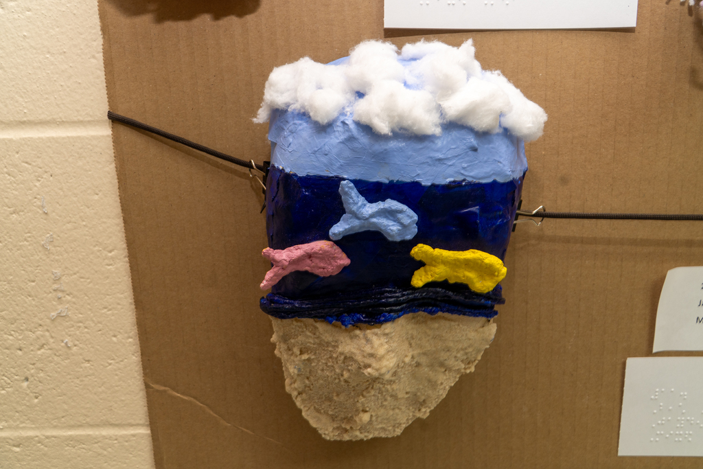 A mask created by a student that looks like a beach with tan color sand, blue for the water, white cotton balls for the clouds and creation of fish swimming through the blue water held for display on a cardboard. 