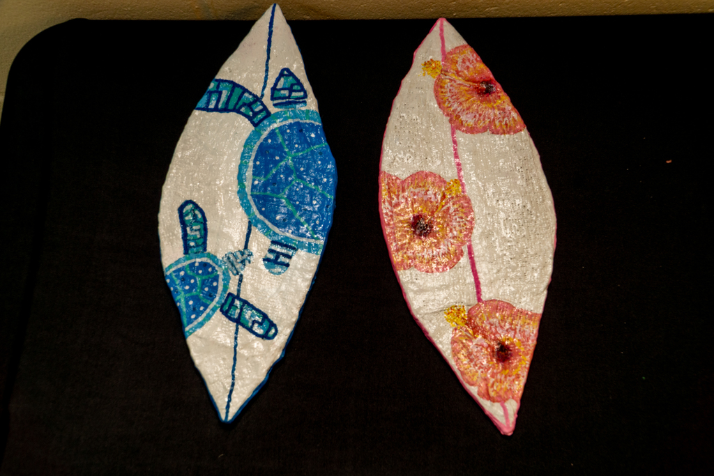  Two clay surfboards are displayed. The left surfboard is white with blue turtles painted on it, while the right surfboard is white decorated with pink, orange, and yellow hibiscus flowers.