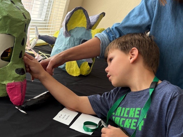 An elementary student inspects a large, green paper mask with large eyes and large pink tongue. A blue and yellow “fish cat” mask in the background has cat ears and fins on the forehead and cheeks. 