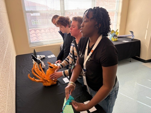 Middle school students are exploring two sculptures with their hands. One is a teal dragon with green wings and the other is an orange, nine-tailed fox.