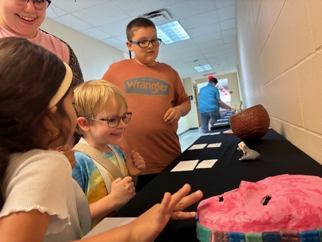 An elementary student with blond hair and glasses smiles with delight at the small sculpture of gray rat wearing a white hat. Another student is feeling the textured tiles that line the edge of pink mask. 