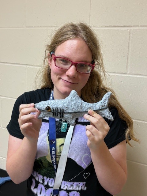 Kaitlyn is smiling while holding up her paper mache shark sculpture, JAWS. It is painted gray, and black yarn forms a stripe and gills. A glass marble bead represents the eye. 