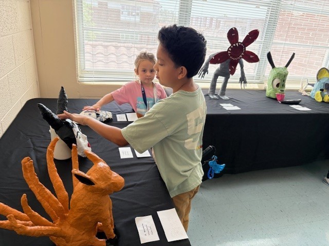 Elementary students explore a Maleficent mask with large black horns. An orange, nine-tailed fox is in the foreground. A Demogorgon with a red, flower-shaped head full of teeth is in the background.