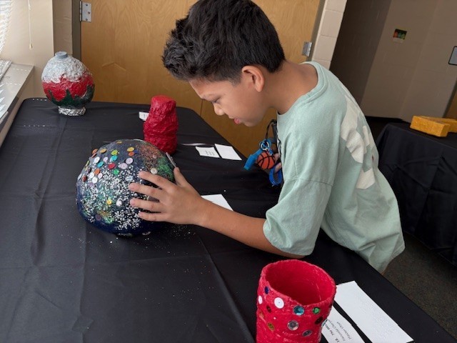 Byran inspects a dark blue, dome-shaped sculpture made of plaster cloth. His is leaning in to feel the sequins and glitter on the surface of piece.
