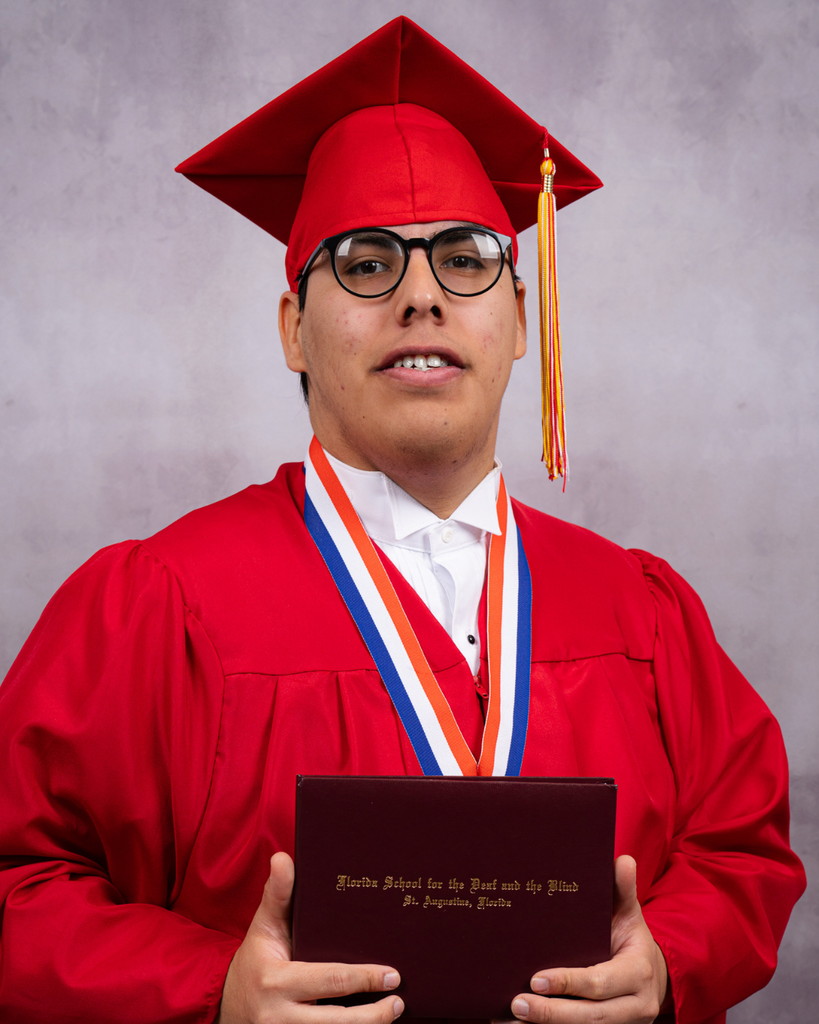 Juan is wearing his red cap and gown smiles for a photo while holding the diploma. 