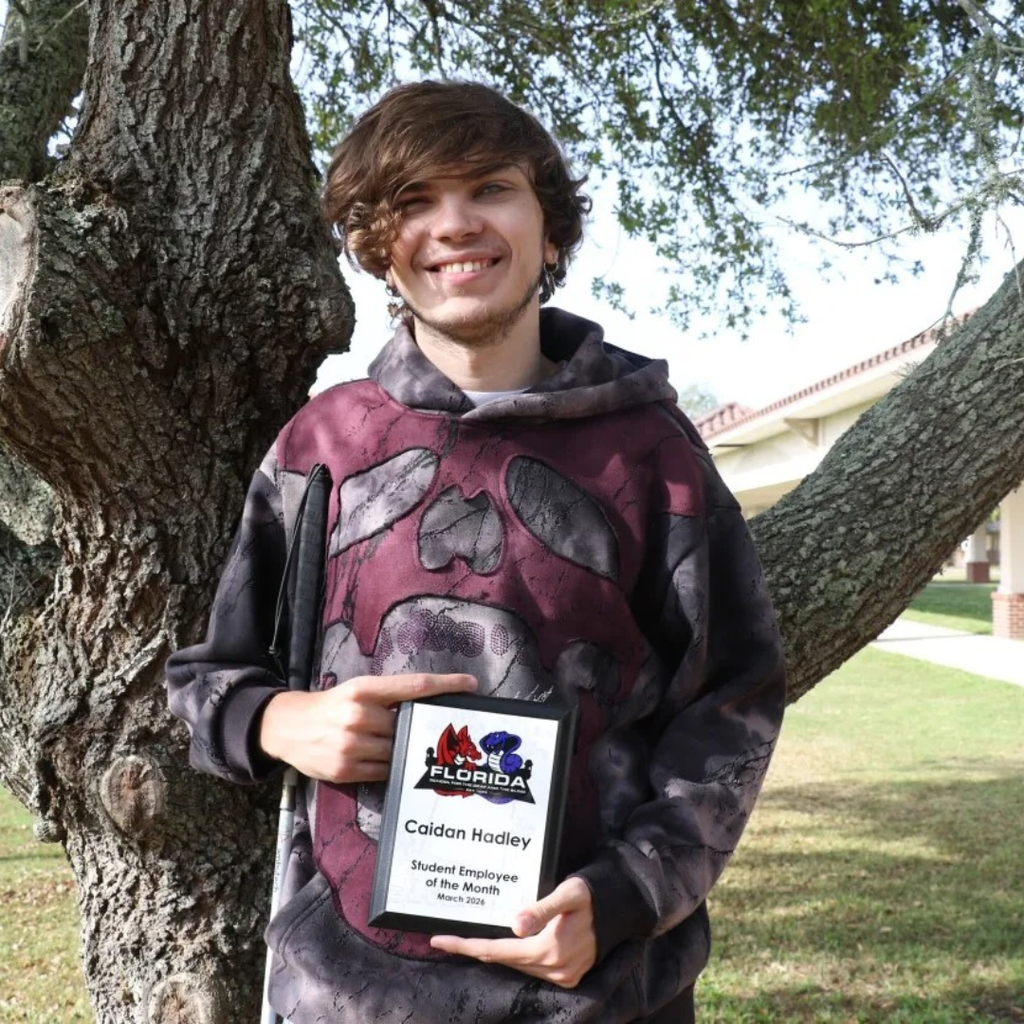 Cadien is holding his "Student Employee of the Month" award smiling by a tree outside. 