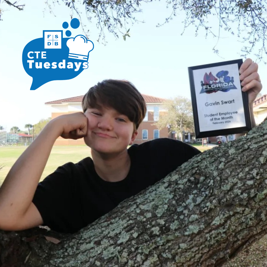 Gavin is holding his "Student Employee of the Month" award smiling for his photo by a tree outside. 