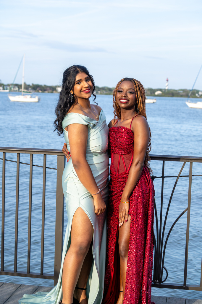 Two friends dressed in green and red prom dresses smile for a photo outside of River House by the water. 