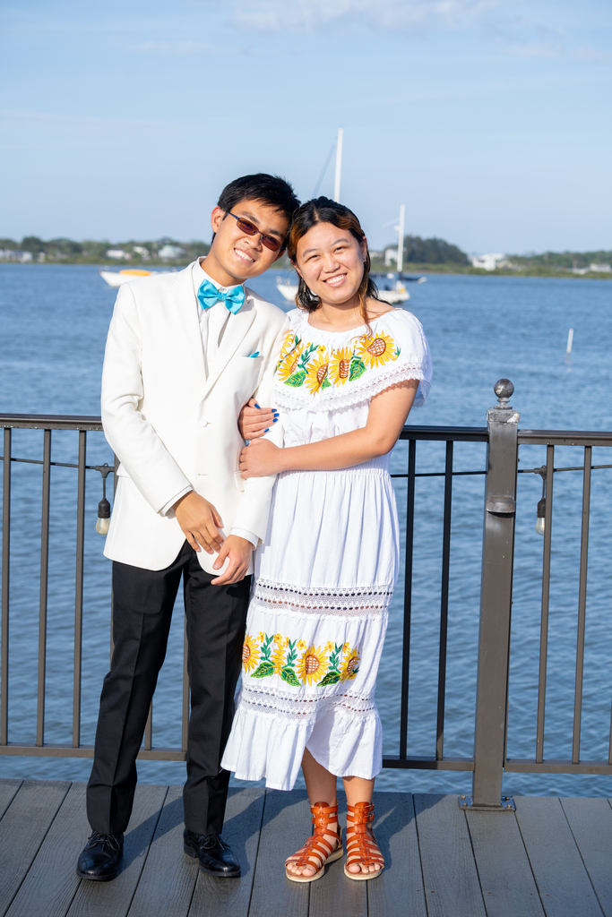 A prom date couple dressed in white smile for a photo outside of the River House by the water. 