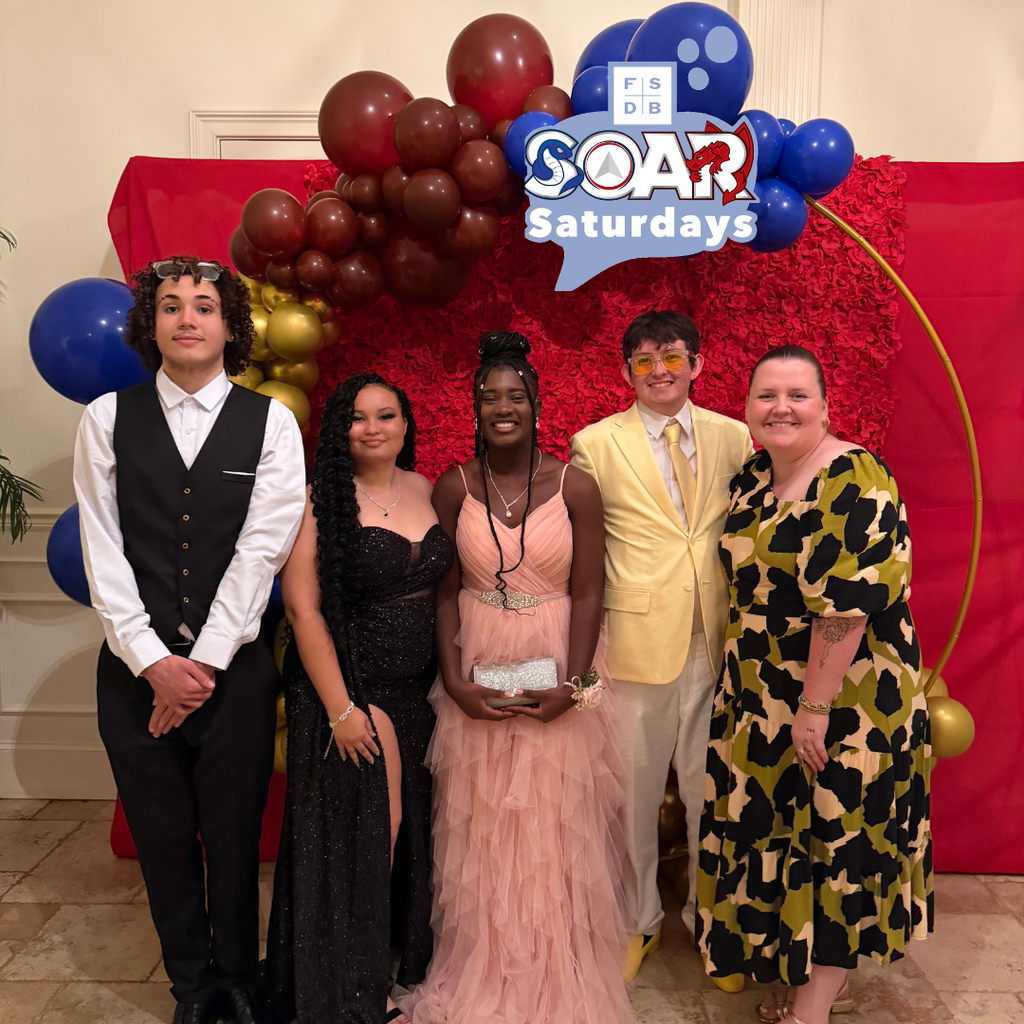 Group of Soar students smile with their SOAR teacher in front of a prom backdrop inside the River House. 