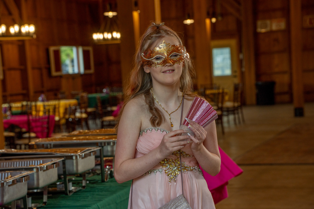 A student is wearing a pink dress and gold masks smiles inside the barn.